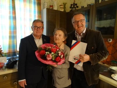 Drei Menschen stehen zusammen in einem Raum, die Frau hält einen Blumenstrauß und der Mann rechts hält ein Zertifikat.