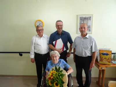 Eine ältere Frau im Rollstuhl, umgeben von drei lächelnden Männern, einer hält einen Blumenstrauß und ein anderer ein Dokument. Der Hintergrund zeigt eine Wand mit einer Uhr und einem gerahmten Bild.