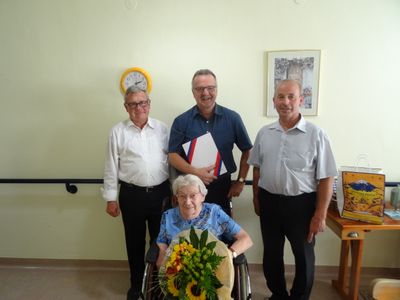 Eine ältere Frau im Rollstuhl, umgeben von drei lächelnden Männern, einer hält einen Blumenstrauß und ein anderer ein Dokument. Der Hintergrund zeigt eine Wand mit einer Uhr und einem gerahmten Bild.