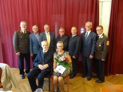 Eine Gruppe von Menschen in formeller Kleidung, einschließlich Militäruniformen, steht und sitzt vor einem roten Vorhang, wobei eine Frau Blumen hält und ein Mann ein Geschenk in der Hand hat.