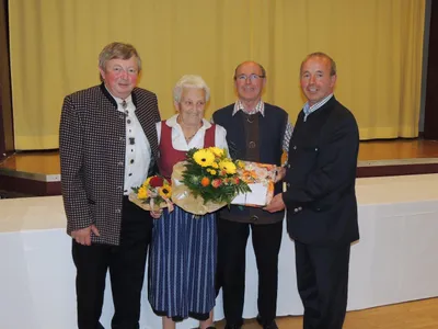 Vier Personen stehen zusammen, zwei Männer und eine Frau, wobei ein Mann einen Blumenstrauß und eine Geschenkbox hält. Die Frau trägt ein traditionelles Kleid.