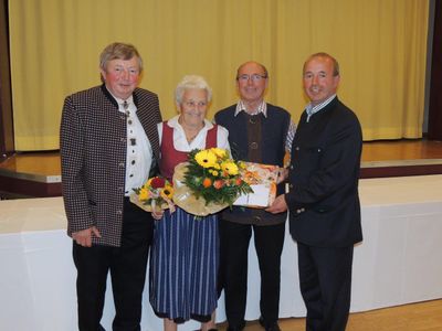 Vier Personen stehen zusammen, zwei Männer und eine Frau, wobei ein Mann einen Blumenstrauß und eine Geschenkbox hält. Die Frau trägt ein traditionelles Kleid.