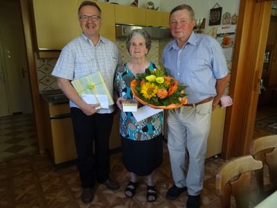 Drei Menschen stehen lächelnd in einer Küche. Ein Mann auf der linken Seite hält ein verpacktes Geschenk, eine Frau in der Mitte hält einen Blumenstrauß und ein Mann auf der rechten Seite trägt einen Gürtel.