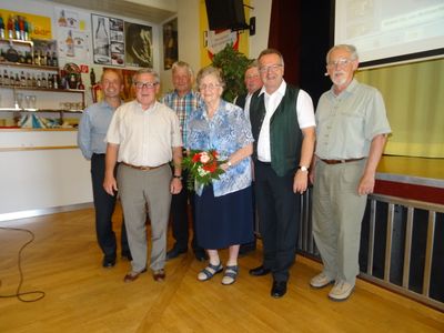 Eine Gruppe von sechs Erwachsenen, darunter eine Frau mit einem Blumenstrauß, steht in einem Raum mit Holzböden und einer dekorativen Pflanze im Hintergrund.