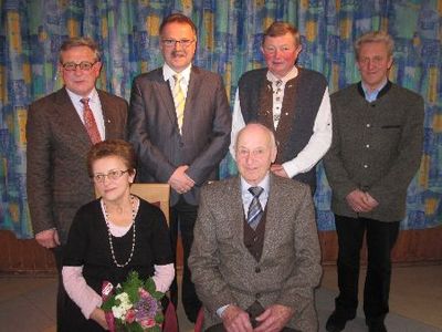 Eine Gruppe von sechs Personen, drei Männer und drei Frauen, steht vor einem farbenfrohen Hintergrund. Der Mann in der Mitte sitzt und lächelt, während die anderen hinter ihm stehen. Die Frau links hält einen Blumenstrauß.