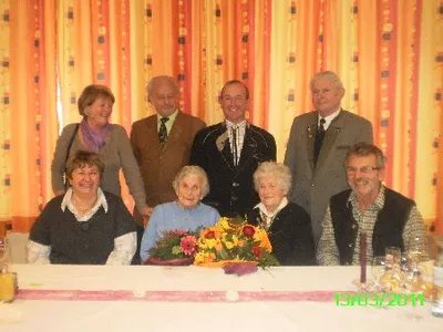 Eine Gruppe älterer Menschen, darunter zwei ältere Frauen, lächeln und posieren für ein Foto an einem Tisch mit Blumen und Getränken.