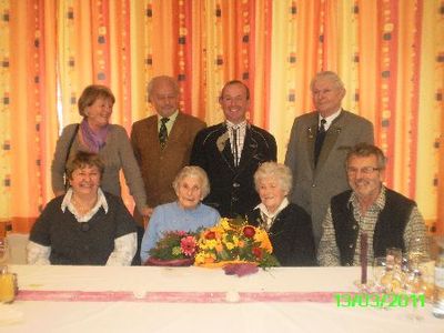 Eine Gruppe älterer Menschen, darunter zwei ältere Frauen, lächeln und posieren für ein Foto an einem Tisch mit Blumen und Getränken.