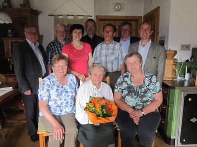 Eine Gruppe von Menschen, darunter eine ältere Frau mit Blumen, versammelt sich in einem Raum für ein Foto. Sie alle lächeln und scheinen für die Kamera zu posieren.