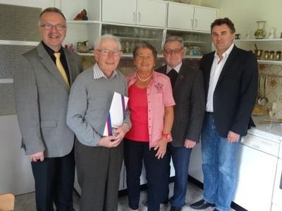 Fünf Personen stehen in einer Küche und posieren für ein Foto. Der Mann in der Mitte hält ein Buch und eine Trophäe. Die Frau neben ihm trägt eine Halskette und ein rosa Oberteil. Der Mann links trägt eine Krawatte. Die anderen beiden Männer tragen Anzüge.