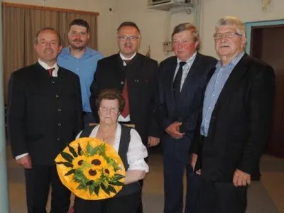 Fünf Männer und eine ältere Frau stehen zusammen und lächeln für ein Foto. Die Frau hält einen Sonnenblumenstrauß. Sie sind formell gekleidet.
