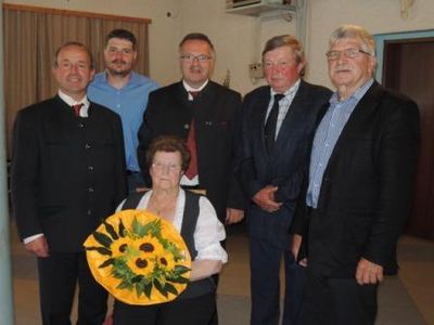 Fünf Männer und eine ältere Frau stehen zusammen und lächeln für ein Foto. Die Frau hält einen Sonnenblumenstrauß. Sie sind formell gekleidet.