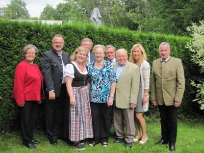 Eine Gruppe von Menschen, darunter Männer und Frauen, steht lächelnd und posiert für ein Foto vor einer Hecke.