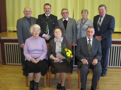 Eine Gruppe älterer Personen, darunter zwei Frauen mit Blumen, sitzen und stehen in einem Raum mit gelbem Vorhang im Hintergrund.