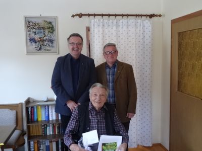 Drei Männer stehen in einem Raum, einer sitzt in einem Stuhl und hält ein Buch. Zwei Männer stehen hinter ihm, einer trägt eine Brille. Ein Bücherregal steht hinter ihnen, und ein Bilderrahmen ist an der Wand montiert.