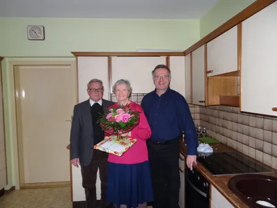 Eine ältere Frau in einem rosa Pullover steht zwischen zwei Männern in einer Küche, hält einen Blumenstrauß und eine Geschenkbox. Der Mann links trägt einen Anzug und eine Krawatte, der Mann rechts ein blaues Hemd. Hinter ihnen steht eine Küchentheke mit Weingläsern und einem Herd.