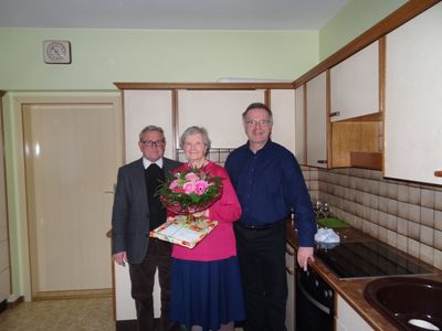 Eine ältere Frau in einem rosa Pullover steht zwischen zwei Männern in einer Küche, hält einen Blumenstrauß und eine Geschenkbox. Der Mann links trägt einen Anzug und eine Krawatte, der Mann rechts ein blaues Hemd. Hinter ihnen steht eine Küchentheke mit Weingläsern und einem Herd.