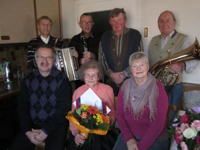 Eine Gruppe älterer Musiker, darunter eine Frau mit Blumen, posiert für ein Foto in einem Raum. Sie halten Musikinstrumente wie Trompete, Akkordeon und Klarinette.