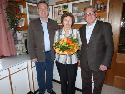 Drei Menschen stehen in einer Küche, eine Frau hält einen Blumenstrauß und zwei Männer stehen auf beiden Seiten von ihr.