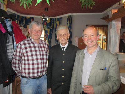 Drei Männer stehen zusammen in einem Raum mit einer roten Decke und blauen Vorhängen. Der Mann auf der linken Seite trägt ein kariertes Hemd, der Mann in der Mitte einen dunklen Anzug und der Mann auf der rechten Seite einen hellgrünen Blazer.