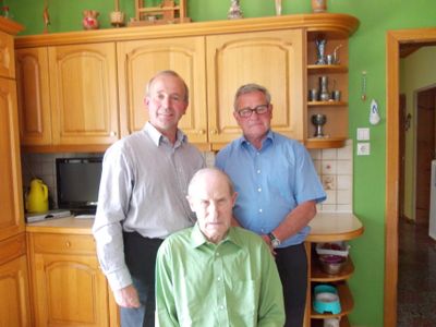 Drei Männer stehen in einer Küche, zwei ältere Männer flankieren einen älteren Mann in einem grünen Hemd. Hinter ihnen sind Holzschränke und Regale mit verschiedenen Gegenständen zu sehen.