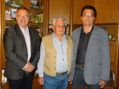 Drei Männer stehen zusammen in einem Raum mit Holzschränken und lächeln für ein Foto. Der Mann links trägt einen dunklen Anzug, der Mann in der Mitte ein hellblaues Hemd und eine beige Weste, und der Mann rechts eine graue Jacke.