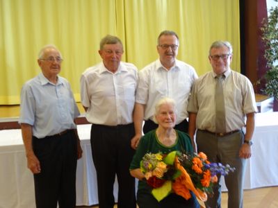 Fünf ältere Männer und eine ältere Frau stehen vor einem gelben Vorhang und lächeln für ein Foto. Die Frau hält einen Blumenstrauß.