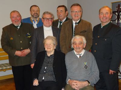 Eine Gruppe älterer Männer und Frauen steht und sitzt in einer formellen Umgebung, möglicherweise für ein Foto. Sie tragen formelle Kleidung.