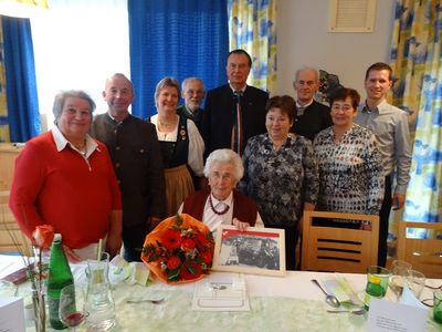 Eine Gruppe von Menschen versammelt sich um einen Tisch, um einen Geburtstag zu feiern. Eine ältere Frau sitzt in der Mitte, hält ein gerahmtes Foto und einen Blumenstrauß. Hinter ihr stehen andere und lächeln, einige tragen traditionelle Kleidung. Der Tisch ist mit Weingläsern, Flaschen und einer Serviette gedeckt.