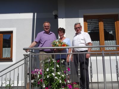 Drei Personen stehen auf einem Balkon und lächeln. Ein Mann in Lila hält einen Blumenstrauß. Eine Frau in Blau steht zwischen zwei Männern, einer in Weiß.