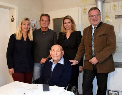 Eine Gruppe von Menschen steht um einen Tisch herum und lächelt für ein Foto. Ein älterer Mann im Rollstuhl steht in der Mitte, mit einem Dokument auf dem Tisch vor ihm.