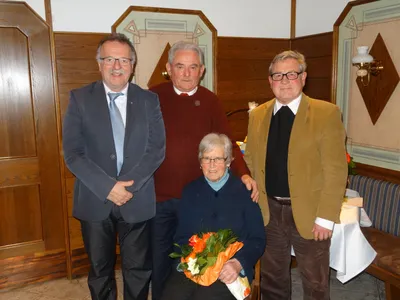 Drei Männer und eine ältere Frau stehen lächelnd zusammen, wobei die Frau einen Blumenstrauß hält. Die Männer sind formell gekleidet, und sie scheinen sich bei einer formellen Veranstaltung zu befinden.