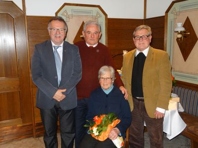 Drei Männer und eine ältere Frau stehen lächelnd zusammen, wobei die Frau einen Blumenstrauß hält. Die Männer sind formell gekleidet, und sie scheinen sich bei einer formellen Veranstaltung zu befinden.
