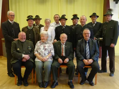 Eine Gruppe von Menschen in traditioneller Kleidung, einschließlich Hüten, sitzt in einer Reihe auf Stühlen und lächelt für ein Foto. Der Ort scheint ein formelles Ereignis zu sein.