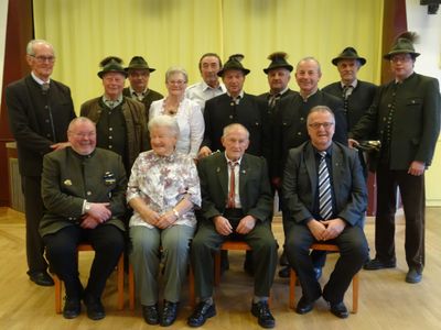 Eine Gruppe von Menschen in traditioneller Kleidung, einschließlich Hüten, sitzt in einer Reihe auf Stühlen und lächelt für ein Foto. Der Ort scheint ein formelles Ereignis zu sein.