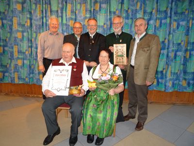 Eine Gruppe von Menschen steht lächelnd vor einem Vorhang. Ein Mann und eine Frau sitzen auf einem Stuhl und halten einen Blumenstrauß und ein Zertifikat. Der Mann hält einen Korb. Dahinter stehen andere, einer hält eine Holztafel.