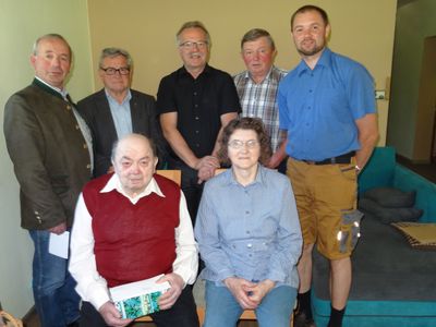 Eine Gruppe von sieben Personen, darunter ältere Männer und Frauen, stehen und sitzen in einem Raum und lächeln für ein Foto.