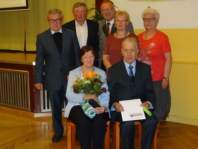Eine Gruppe älterer Menschen steht und sitzt in einem Raum, wahrscheinlich für eine formelle Veranstaltung. Eine Frau hält einen Blumenstrauß und ein Mann ein Zertifikat.