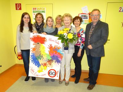 Eine Gruppe von Menschen steht in einem Raum mit einer gelben Wand, hält ein farbenfrohes Poster und einen Blumenstrauß. Das Poster sagt 'Alles Gute' und '60'. Die Frau, die das Poster hält, lächelt.