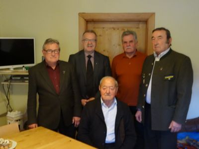 Fünf Männer in formeller Kleidung stehen in einem Raum, einer sitzt in der Mitte. Sie scheinen für ein Foto zu posieren. Hinter ihnen ist eine Holztür und ein Monitor auf der linken Seite.