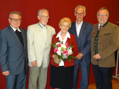 Fünf ältere Personen stehen vor einem roten Hintergrund und lächeln für ein Foto. Die Frau in der Mitte hält einen Blumenstrauß.