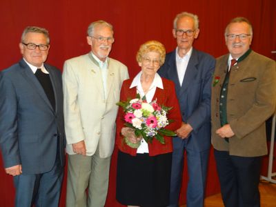 Fünf ältere Personen stehen vor einem roten Hintergrund und lächeln für ein Foto. Die Frau in der Mitte hält einen Blumenstrauß.