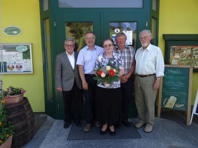 Fünf Personen stehen vor einem gelben Gebäude mit einer grünen Tür und lächeln für ein Foto. Die Frau in der Mitte hält einen Blumenstrauß.