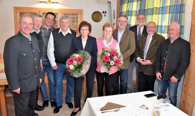 Eine Gruppe älterer Menschen, zwei Frauen mit Blumensträußen, stehen in einem Raum mit einem Tisch und einem Vorhang im Hintergrund.