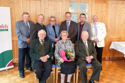 Eine Gruppe älterer Männer und Frauen, in formeller Kleidung gekleidet, versammelt sich in einem Raum. Eine Frau in der Mitte hält einen Blumenstrauß. Sie alle lächeln und scheinen für ein Foto zu posieren.