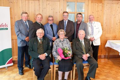 Eine Gruppe älterer Männer und Frauen, in formeller Kleidung gekleidet, versammelt sich in einem Raum. Eine Frau in der Mitte hält einen Blumenstrauß. Sie alle lächeln und scheinen für ein Foto zu posieren.