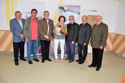 Eine Gruppe von Menschen in formeller Kleidung steht vor einem Banner. Eine Frau in der Mitte hält einen Blumenstrauß.