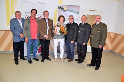 Eine Gruppe von Menschen in formeller Kleidung steht vor einem Banner. Eine Frau in der Mitte hält einen Blumenstrauß.
