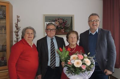 Vier Personen, zwei Männer und zwei Frauen, stehen eng beieinander in einem Raum. Die Frau auf der rechten Seite hält einen Blumenstrauß. Hinter ihnen befindet sich ein Blumenbild an der Wand.