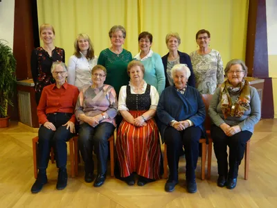 Eine Gruppe älterer Frauen in verschiedenen Outfits sitzt vor einem gelben Vorhang auf Stühlen.