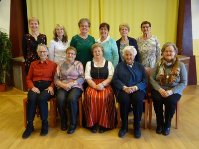 Eine Gruppe älterer Frauen in verschiedenen Outfits sitzt vor einem gelben Vorhang auf Stühlen.